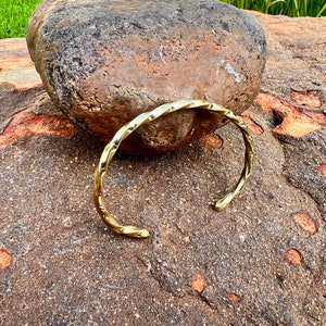 Könnte beinhalten: Ein goldfarbenes, gedrehtes Metall-Armreif-Armband. Das Armband liegt auf einem rauen, braunen Stein.