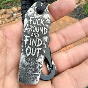 May include: A black paracord keyring featuring a rectangular metal tag with a distressed silver finish. The tag is engraved with the text FUCK AROUND AND FIND OUT in a stylised font, surrounded by skull imagery, and includes a black metal carabiner.