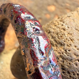 May include: Close-up of a handcrafted, curved metal object with a textured surface. The metal is painted with a mix of red, silver, and black colors, creating a unique, artistic appearance. The object is set against a blurred, natural background.