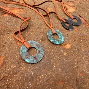 May include: Three leather cord necklaces with circular metal pendants. The pendants are different sizes and have a rustic, aged look. The largest pendant has an inscription in an unknown language.