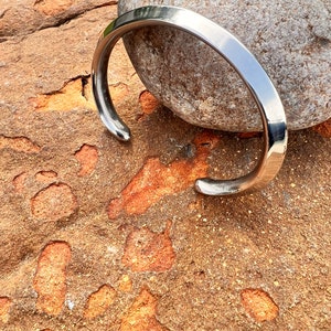 Puede incluir: Un brazalete de plata con un acabado liso y pulido. El brazalete está abierto y tiene un diseño simple y minimalista.