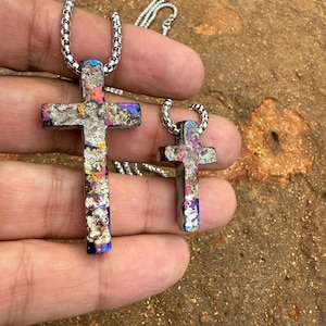 May include: Two silver-toned cross pendants with colorful iridescent accents. The crosses are suspended from matching chain necklaces. The larger cross is approximately 2 inches long, while the smaller one is about 1 inch long. The background is a textured, earthy surface.