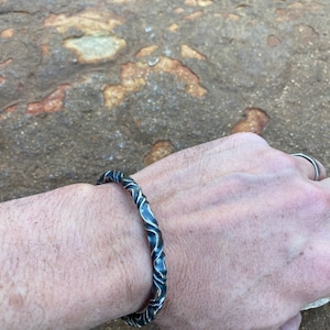 Puede incluir: Un brazalete de plata con un diseño texturizado y ondulado. El brazalete está hecho de metal y tiene un acabado pulido.