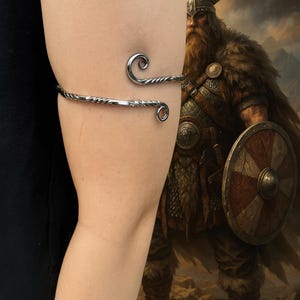 Puede incluir: Un brazalete plateado con un diseño retorcido y extremos rizados. El brazalete se envuelve alrededor del brazo, creando un accesorio decorativo. El fondo presenta una pintura de un guerrero vikingo.