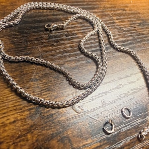 May include: A silver-colored chain necklace with a lobster clasp, lying on a wooden surface. The necklace is made of small, interconnected links. Several small metal components are also present, including a clasp and jump rings.