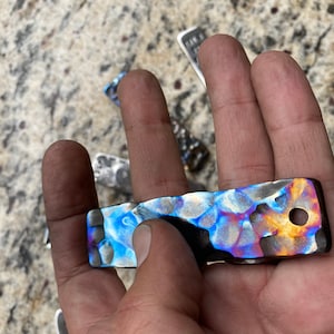 May include: A rectangular metal object with a hammered texture and a hole. The object has a vibrant color scheme, with hues of blue, purple, and gold. Other similar objects are visible in the background.