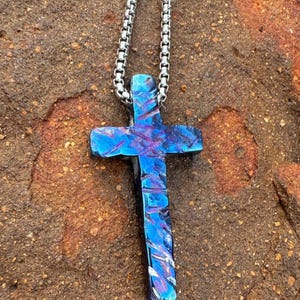 May include: A silver chain necklace with a blue and purple metal cross pendant. The cross has a textured surface and is a unique piece of jewelry.