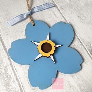 May include: A blue wooden forget-me-not flower with a yellow centre and white star-shaped accents. The flower is hanging from a blue and white gingham ribbon.