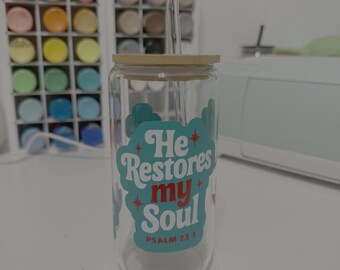 Puede incluir: Un vaso de vidrio transparente con tapa de madera y pajita de vidrio curvada. El vaso presenta un gráfico turquesa y rojo con el texto "He Restores my Soul" y "Psalm 23:3".