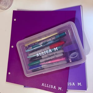 May include: A clear plastic pencil case filled with pens and markers, resting on top of two purple folders. The text "ALLISA M" is printed on the folders and the pens. A small purple container of Crayola ink is also visible.