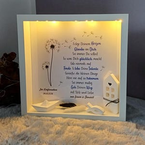 Könnte beinhalten: Ein weiß gerahmter Schattenkasten mit einer gedruckten Botschaft und dekorativen Elementen. Die Botschaft enthält Text und Pusteblumen-Illustrationen. Die Box enthält ein Papierboot, eine Feder, einen Fisch und ein kleines Haus. Das Datum 24.03.2024 ist sichtbar.