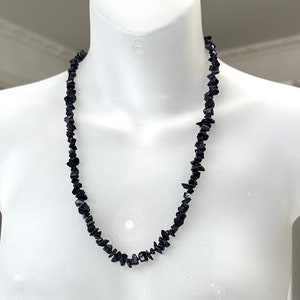 May include: A black gemstone necklace displayed on a white mannequin. The necklace is made of small, irregularly shaped black stones. The mannequin is on a clear, round base.
