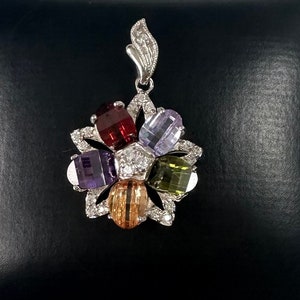 May include: A silver pendant with a flower design featuring six colorful gemstones. The gemstones are red, purple, green, orange, and two shades of purple.