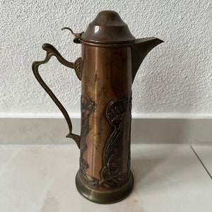 May include: A copper pitcher with an ornate design. The pitcher has a lid and a handle. The pitcher is decorated with a floral pattern.