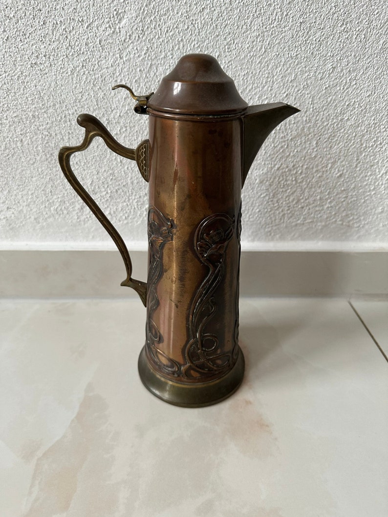 May include: A copper pitcher with an ornate design. The pitcher has a lid and a handle. The pitcher is decorated with a floral pattern.
