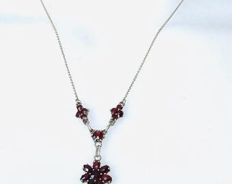 May include: A silver necklace with a floral design. The necklace has a delicate chain and three clusters of dark red gemstones. The main feature is a larger, flower-shaped pendant with multiple red gemstones.