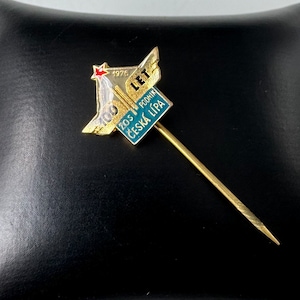 May include: A gold-coloured pin with a blue and green design. The pin features a rocket ship with the text "1976 100 LET ZOS PODNIK CESKA LIPA" on it.