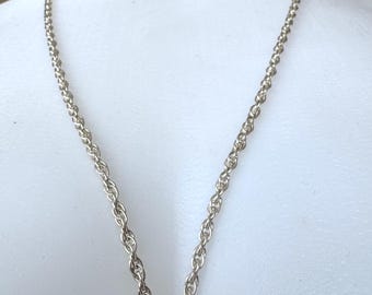 May include: A silver-colored necklace with a rope chain design. The chain is draped around a white mannequin, showcasing the necklace's shape and style. The chain is made of small, tightly linked segments, creating a textured appearance.