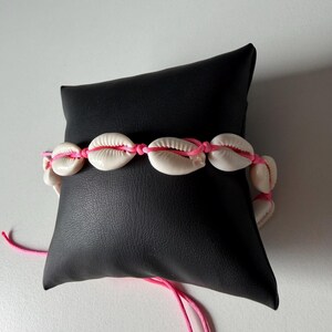May include: A seashell bracelet featuring white shells strung on a bright pink cord. The bracelet is displayed on a black surface. The shells are oval-shaped and have a natural, textured appearance. The pink cord adds a pop of color.