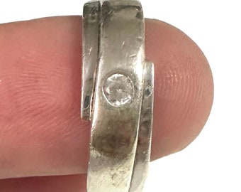 Puede incluir: Un anillo de tono plateado con una banda de estilo bypass y un acabado liso y pulido. El centro de la banda está engastado con una sola piedra redonda, transparente y facetada, creando un aspecto limpio y reflectante en la superficie.
