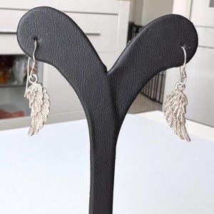 Pu&ograve; includere: Orecchini a forma di piuma color argento esposti su un espositore nero per gioielli. Gli orecchini presentano un design dettagliato ad ala, con texture di piume visibili. I ganci sono semplici. Lo sfondo &egrave; un interno sfocato.