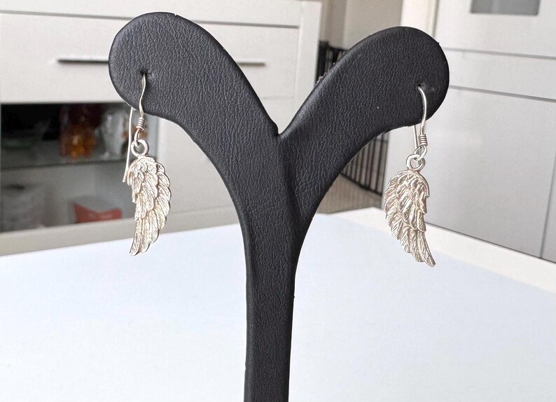 Pu&ograve; includere: Orecchini a forma di piuma color argento esposti su un espositore nero per gioielli. Gli orecchini presentano un design dettagliato ad ala, con texture di piume visibili. I ganci sono semplici. Lo sfondo &egrave; un interno sfocato.