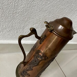 May include: A copper and brass pitcher with an ornate design. The pitcher has a hinged lid and a handle.