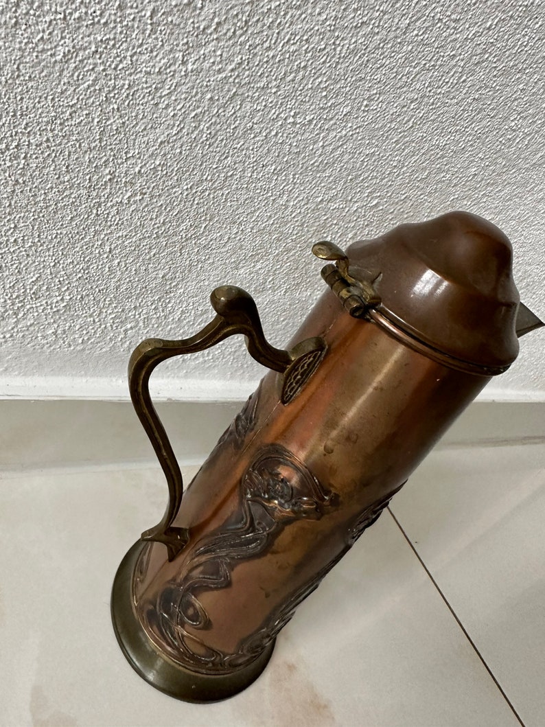 May include: A copper and brass pitcher with an ornate design. The pitcher has a hinged lid and a handle.