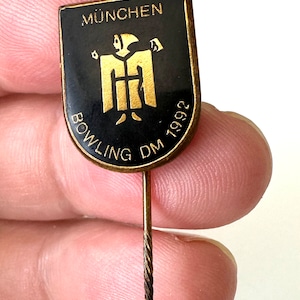 Könnte beinhalten: Eine goldene und schwarze Anstecknadel mit dem Text "MÜNCHEN BOWLING DM 1992" und einer stilisierten Figur einer Person in einer Robe.