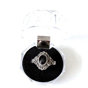 Op de afbeelding: Een zilveren ring met een ovale zwarte steen in het midden, omringd door kleine, ronde accenten. De ring wordt gepresenteerd in een transparante, scharnierende sieradendoos met een zwart fluwelen interieur.