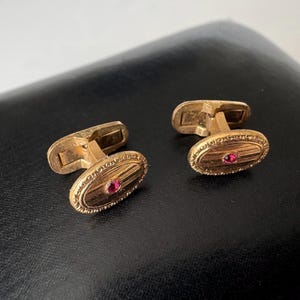 May include: A pair of gold-toned cufflinks with an oval design. Each cufflink features a small, round, red gemstone in the center and an engraved border. The cufflinks are displayed on a dark surface.