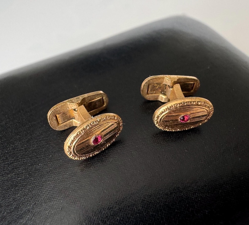 May include: A pair of gold-toned cufflinks with an oval design. Each cufflink features a small, round, red gemstone in the center and an engraved border. The cufflinks are displayed on a dark surface.