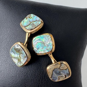 May include: Gold-toned cufflinks with iridescent abalone shell inlays. The square-shaped inlays display a range of colors, including blues, greens, and hints of gold. The cufflinks are set against a black background.