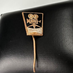 May include: A gold-colored pin with a black and white film camera design. The camera has the letters "FS" on it. The text "BARRANDOV" is written on a banner below the camera.