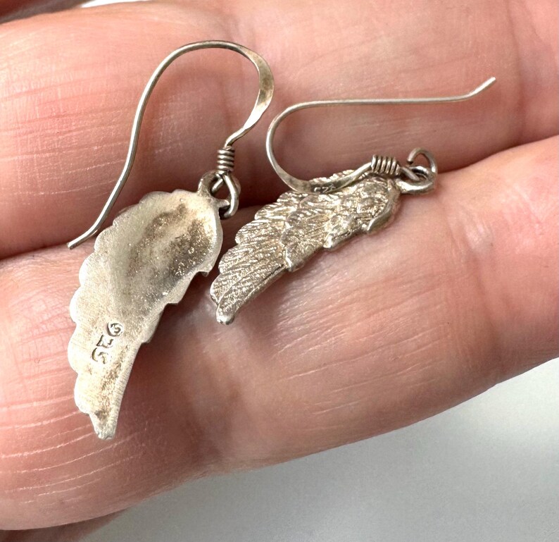 Pu&ograve; includere: Un paio di orecchini color argento, ciascuno con un ciondolo a forma di ala. I ciondoli hanno un design dettagliato a forma di piuma e sono sospesi a un semplice gancio. Gli orecchini sono mostrati su uno sfondo neutro.