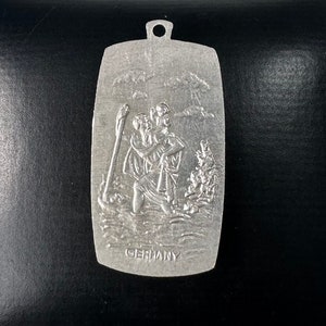 May include: Silver metal pendant featuring a relief sculpture of Saint Christopher carrying a child. The pendant is rectangular with rounded corners and has the word "GERMANY" inscribed at the bottom.