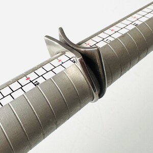 May include: A silver ring sizing tool with a ring placed on it. The tool has a ruler with markings in inches.