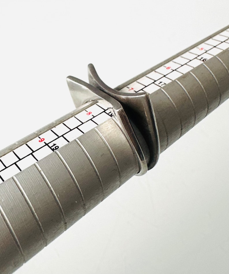 May include: A silver ring sizing tool with a ring placed on it. The tool has a ruler with markings in inches.