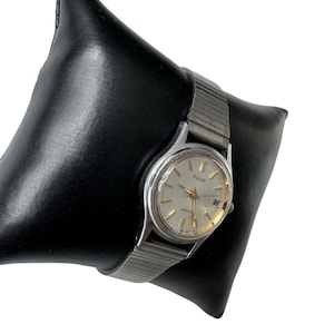 Op de afbeelding: Een zilverkleurig Pulsar quartz horloge met een ronde wijzerplaat in een lichte kleur, goudkleurige uurmarkeringen en een datumvenster op de drie-uurpositie. Het horloge heeft een flexibele, rekbare metalen schakelarmband.