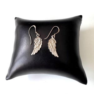 Pu&ograve; includere: Un paio di orecchini a forma di ala color argento con chiusura a gancio. Gli orecchini sono esposti su un cuscino di pelle nera. Le ali presentano dettagliati disegni di piume.