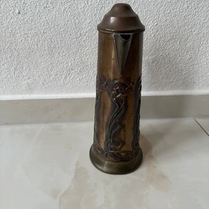May include: A tall, copper pitcher with an ornate, embossed design. The pitcher has a silver spout and a lid.