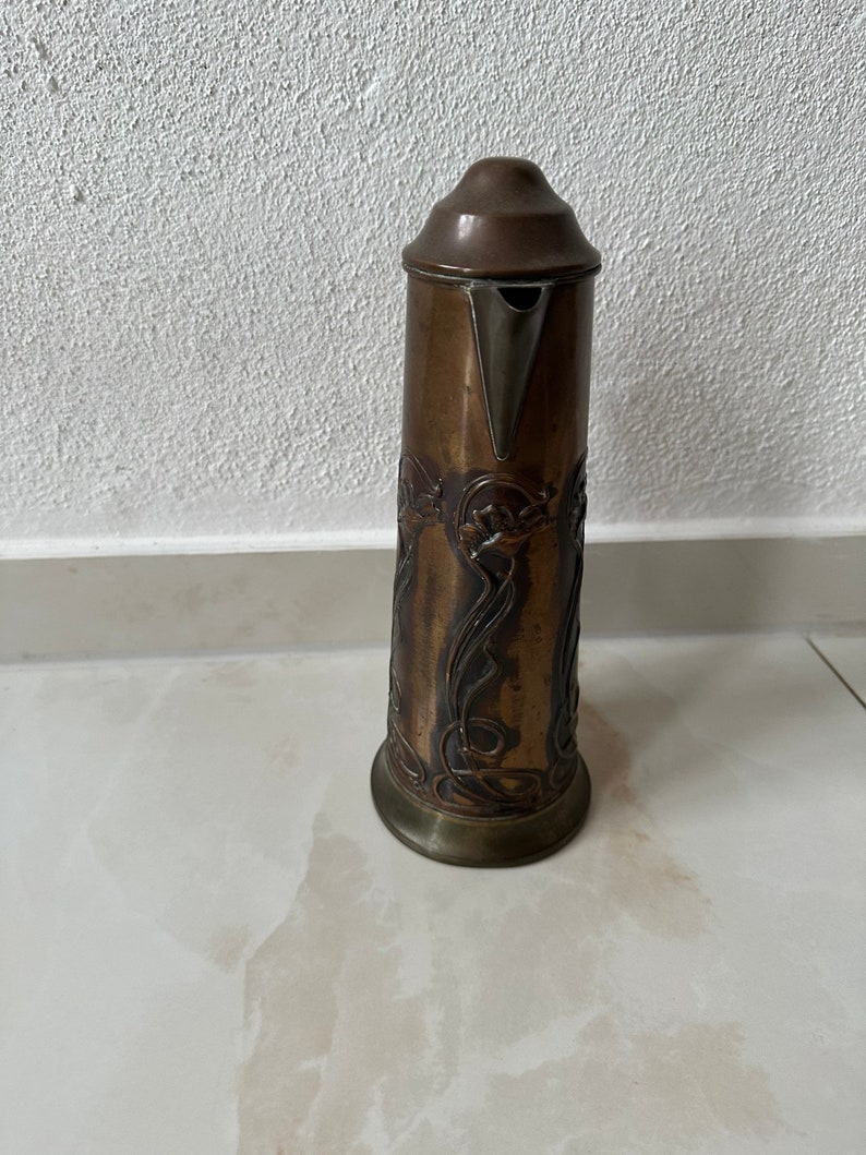 May include: A tall, copper pitcher with an ornate, embossed design. The pitcher has a silver spout and a lid.