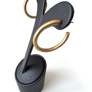 May include: Gold-colored hoop earrings displayed on a black earring stand. The earrings are a simple, circular design. The stand is black with a rounded base and a vertical post with two earring holders. The background is white.