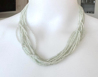 May include: A multi-strand necklace composed of small, pale green beads. The necklace is draped around a white mannequin's neck, showcasing its design. The strands are loosely twisted together, creating a textured appearance. The beads reflect light, giving a subtle shimmer.