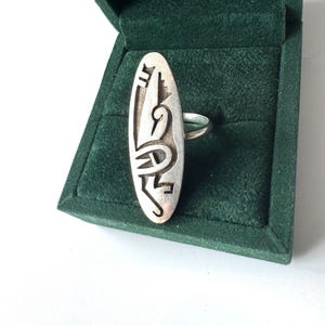 Könnte beinhalten: Silberfarbener Ring mit länglicher, ovaler Oberfläche und abstraktem Design. Der Ring liegt in einer dunkelgrünen Samt-Schmuckschatulle. Das Design umfasst geometrische und geschwungene Formen, die einen einzigartigen, künstlerischen Look erzeugen.