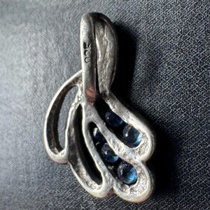 Op de afbeelding: Zilveren hanger met een delicaat, abstract ontwerp met drie rijen blauwe edelstenen. De hanger heeft een lus aan de bovenkant om een ​​ketting aan te bevestigen. De achterkant van de hanger is gestempeld met het nummer "925", wat aangeeft dat hij van sterling zilver is.
