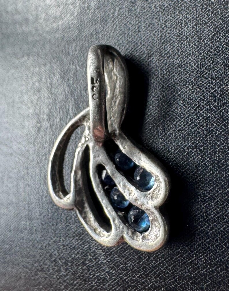Op de afbeelding: Zilveren hanger met een delicaat, abstract ontwerp met drie rijen blauwe edelstenen. De hanger heeft een lus aan de bovenkant om een ​​ketting aan te bevestigen. De achterkant van de hanger is gestempeld met het nummer "925", wat aangeeft dat hij van sterling zilver is.