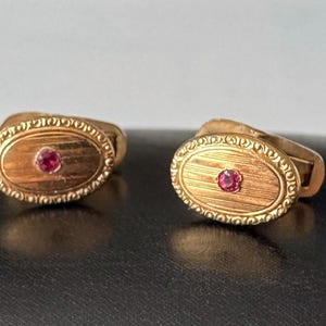 May include: A pair of gold cufflinks with an oval design. Each cufflink features a small, round, red gemstone in the center and a decorative border. The cufflinks are displayed on a dark surface.