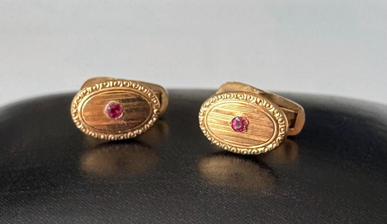 May include: A pair of gold cufflinks with an oval design. Each cufflink features a small, round, red gemstone in the center and a decorative border. The cufflinks are displayed on a dark surface.