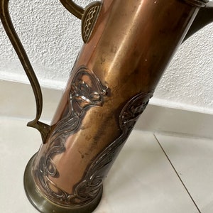 May include: A copper pitcher with an ornate design of stylized flowers and leaves. The pitcher has a brass handle and a brass base.
